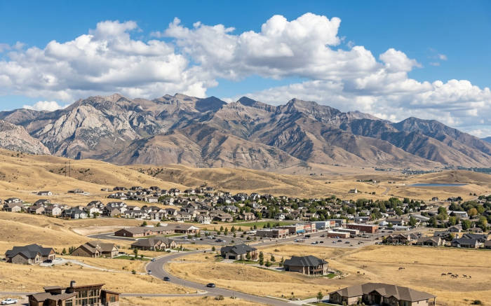 Suburban sprawl in a valley before vast, rugged mountains under a blue, cloudy sky.