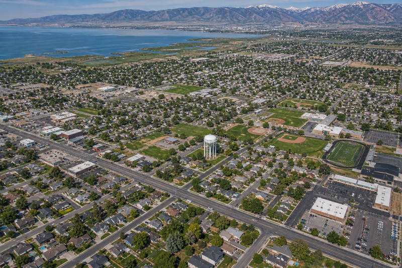 Aerial view: suburban town, water tower, sports fields, houses, lake, and snowy mountains.