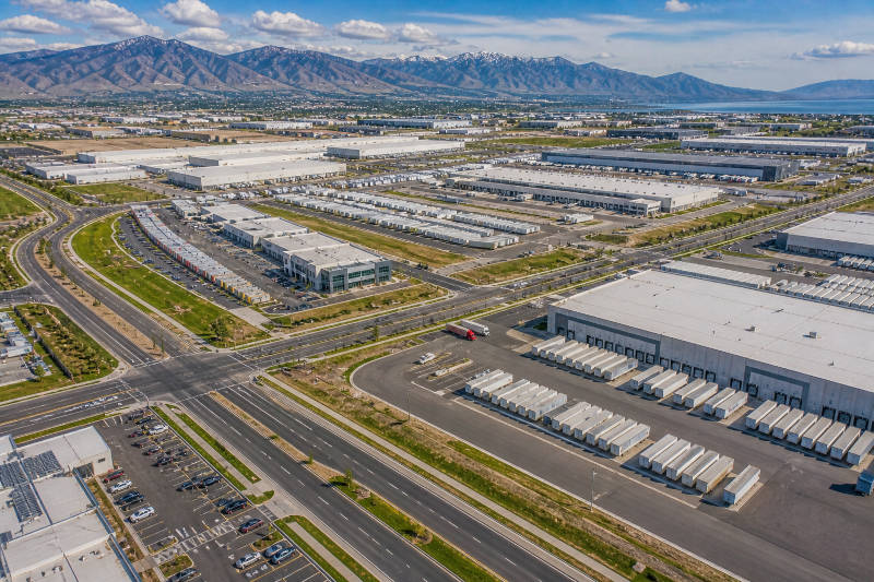 Aerial: vast industrial park, many warehouses, roads, distant snow-capped mountains, and a lake.
