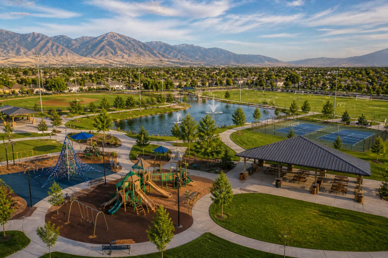 Expansive park features playgrounds, a pond with fountains, sports fields, and distant mountains.